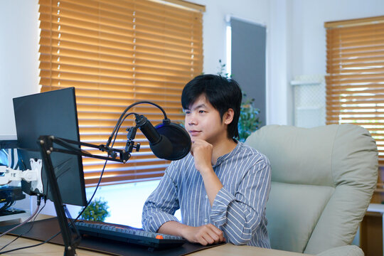 Young Asian blogger man checking script on computer before recording podcast for his social media with a condenser microphone, ideal for remote work, podcasting, or streaming concepts
