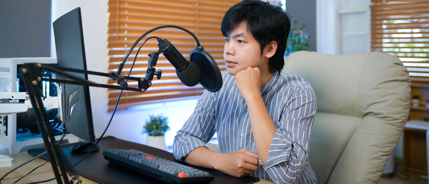 Young Asian blogger man checking script on computer before recording podcast for his social media with a condenser microphone, ideal for remote work, podcasting, or streaming concepts