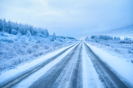 Straight icy snow-covered road with tire tracks through frozen pine forest in Iceland winter. Scenic arctic highway road trip and hazardous driving condition.