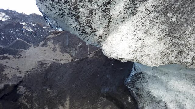 Close-up of a large block of melting glacier ice streaked with volcanic ash, water dripping from its edge, with dark mountains behind, Vatnajokull, Iceland.