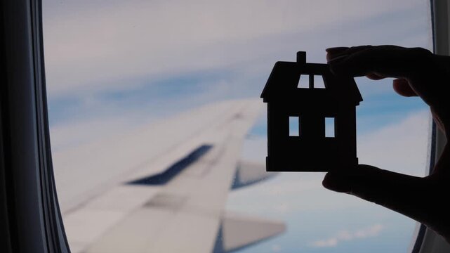 Person hand holding small wooden house model near airplane window during flight.
