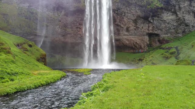 Seljalandsfoss waterfall, Iceland, feeding a small river that winds through bright green grass toward the camera, with mist rising at the foot of the cliff.