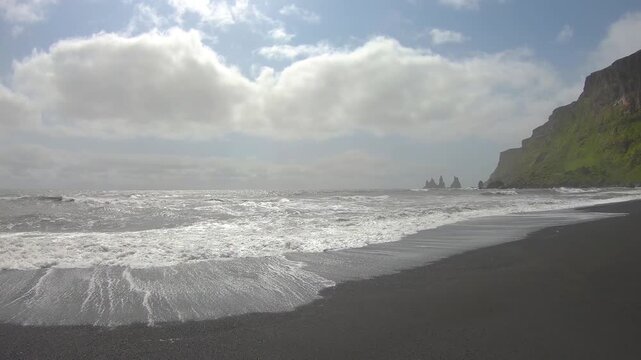 Waves breaking on Reynisfjara black sand beach, with the Reynisdrangar sea stacks and green Reynisfjall cliff in the distance under white clouds.