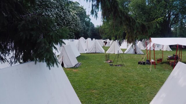 Establishing shot of a historic reenactment camp with numerous white tents pitched on a lush green lawn surrounded by trees dynamic cinematic footage.