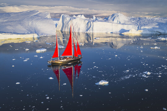 Aerial view of a sailboat with striking red sails navigating through calm waters filled with floating icebergs near the Ilulissat Icefjord in Ilulissat, Avannaata Municipalit, Greenland.