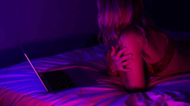 Sensual young woman in lingerie using laptop on bed, online chat and intimate webcam mood in neon night light