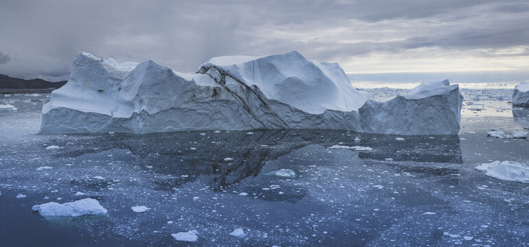Aerial view of a massive iceberg floating in the dark waters of the Ilulissat Icefjord surrounded by scattered ice fragments under a cloudy sky in Ilulissat, Avannaata Municipalit, Greenland.
