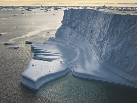 Aerial view of the massive Ilulissat Icefjord iceberg floating in the cold Arctic waters with textured ice surfaces and calm sea under soft light in Ilulissat, Avannaata Kommunia, Greenland.