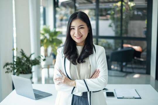 Confident Asian businesswoman smiling with crossed arms near desk while working with laptop inside modern corporate office background for successful professional leader concept