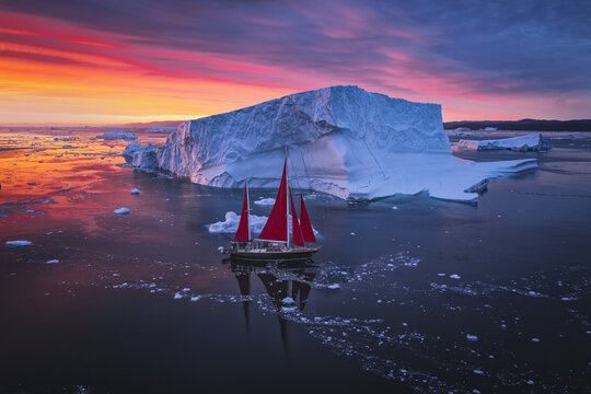 Aerial view of a sailboat with striking red sails navigating through icy waters near a massive iceberg under a vibrant sunset sky in Ilulissat, Avannaata Municipalit, Greenland.