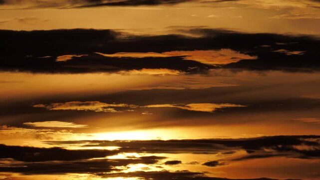 Passing moment of clarity amidst looming shadows and transformation. Drifting horizontal stratus cloud bands slide across sky, casting rich dramatic shadows while obscured sun descends in background.
