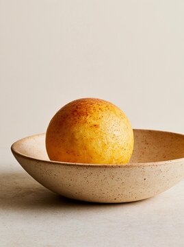Single orange in a small bowl. the bowl is made of a light-colored material with a speckled texture. the orange is round and has a yellowish-orange color with darker spots on its surface.