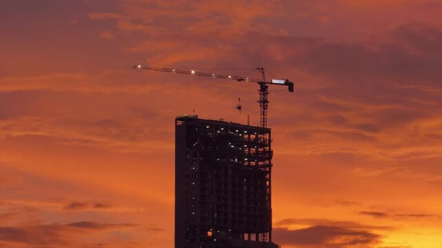 Raw concrete skeleton of residential tower, standing as static monolith while tower crane rotates. Real spectacle unfolds in background canvas, where time-lapse shows dramatic explosion of sunset