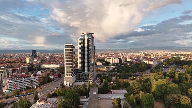 A drone passes beside modern glass high-rise towers while revealing a dense urban skyline and distant mountains.