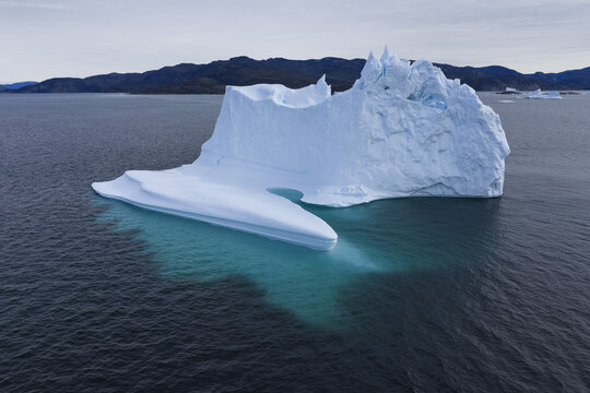 Aerial view of a massive iceberg floating in the deep blue waters of Ilulissat Icefjord with turquoise submerged ice visible beneath the surface in Ilulissat, Avannaata, Greenland.