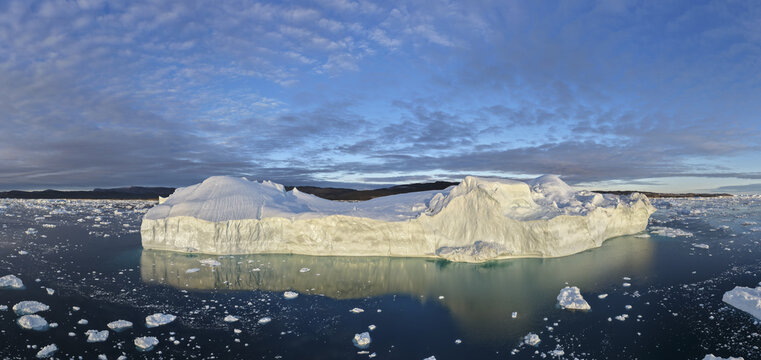 Aerial view of a massive iceberg floating in the Ilulissat Icefjord, surrounded by scattered ice fragments under a vast blue sky in Ilulissat, Avannaata Kommunia, Greenland.