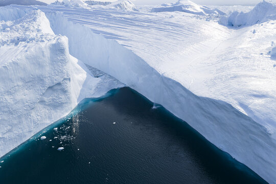 Aerial view of a massive iceberg with towering white ice walls rising above the deep blue water of the Ilulissat Icefjord in Ilulissat, Avannaata Municipalit, Greenland.