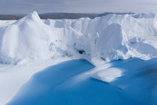 Aerial view of massive icebergs floating in the bright blue waters of the Ilulissat Icefjord, with rugged coastal mountains in the background in Ilulissat, Avannaata, Greenland.
