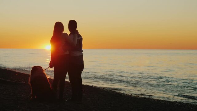 Silhouetted couple embracing on pebble beach during sunset with loyal dog sitting nearby by calm sea waves creating romantic cinematic scene of love and togetherness in beautiful natural environment.