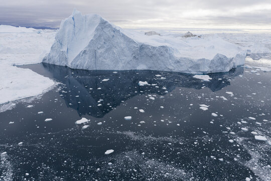 Aerial view of a massive iceberg floating in the dark waters of the Ilulissat Icefjord with scattered sea ice fragments and a cloudy sky in Ilulissat, Avannaata Municipalit, Greenland.