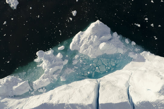 Aerial view of the Ilulissat Icefjord featuring a massive white iceberg with deep cracks floating in dark arctic waters with turquoise hues at the base, Ilulissat, Avannaata Kommunia, Greenland.