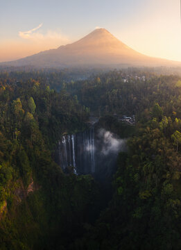 Aerial view of Tumpak Sewu Waterfall surrounded by lush tropical forest with Mount Semeru in the background at sunrise in Kecamatan Pronojiwo, Jawa Timur, Indonesia.