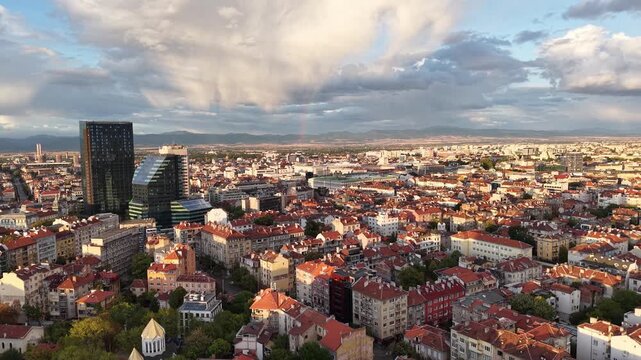 Dense urban neighborhoods with red-tiled roofs and modern high-rises extend toward distant mountains in a moving aerial view.