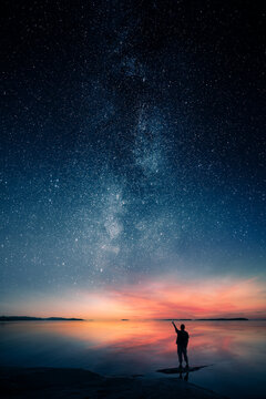 Silhouette of a man pointing at the Milky Way and starry night sky over a calm lake at colorful twilight