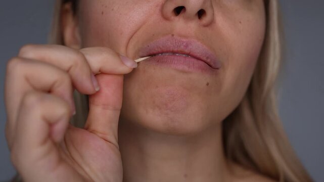Young woman using a wooden toothpick to clean food between teeth, close-up of mouth, lips and hand during dental hygiene routine on gray background
