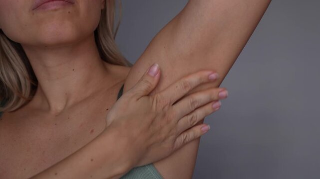 Young woman spreading cream on underarm after shaving, close-up of armpit skincare, moisturizing body lotion, smooth skin and irritation care on gray background