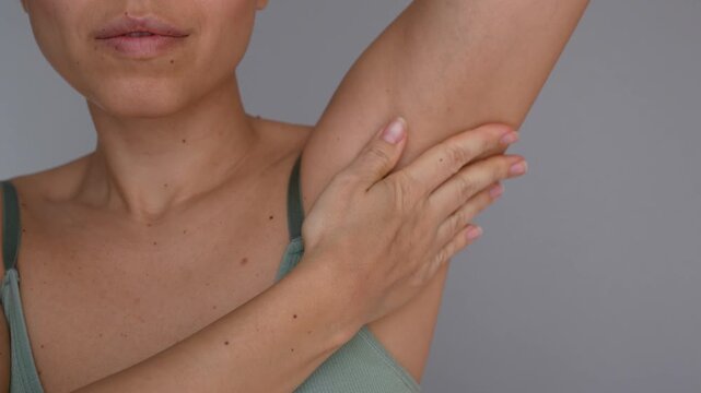 Young woman applying white cream to underarm after shaving, close-up of armpit care, moisturizing lotion, smooth skin and post shaving skincare on gray background