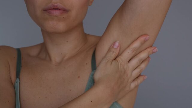 Young woman applying and spreading cream on underarm after shaving, close-up of armpit skincare, smooth hairless skin, irritation care and body hygiene on gray background