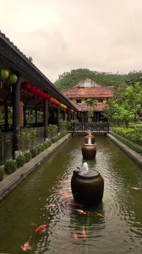 Smooth vertical handheld pan of a traditional Asian garden pond with ornate ceramic urn fountains, koi fish, and hanging lanterns, Ba Na Hills, Da Nang, Vietnam