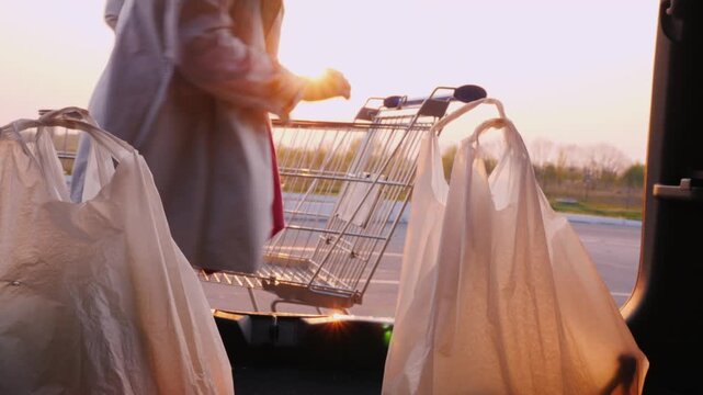 Woman loading groceries from shopping cart into car trunk at sunset, dynamic motion footage.