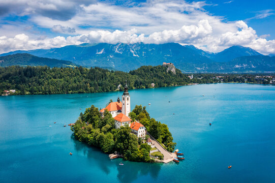 Aerial view of the Pilgrimage Church of the Assumption of Maria on Bled Island surrounded by turquoise water and lush greenery with mountains in the background in Bled, Radovljica, Slovenia.