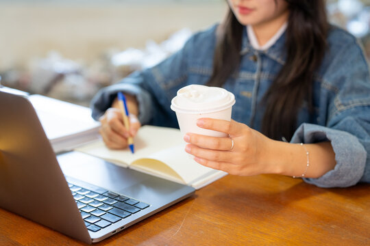 Close up of student hand holding disposable coffee cup and writing on textbook with open laptop screen on desk during study session.