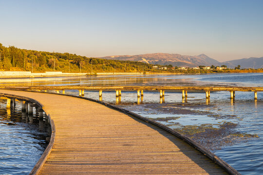 Seaside landscape near the city of Vlore in Albania, Europe. A serene wooden boardwalk curves over tranquil blue lake water toward distant mountains under a clear bright morning sky