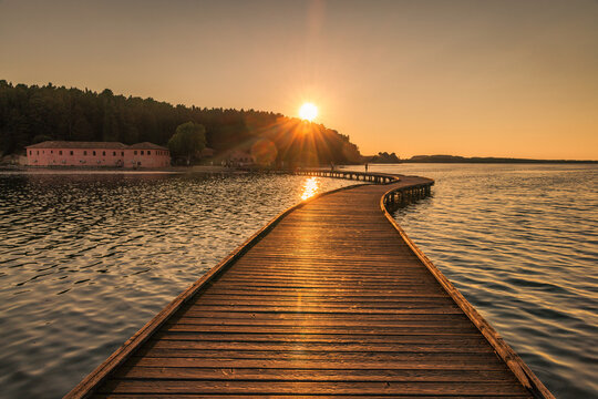 Zvernec Monastery near the city of Vlore in Albania, Europe. A serene wooden boardwalk stretching across calm water toward a warm golden sunset illuminating a distant shoreline peacefully