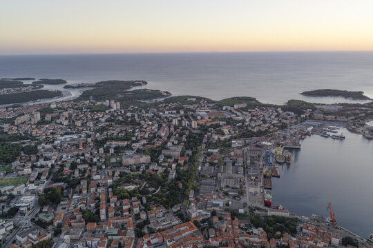 Aerial view of the coastal city of Pula with its historic harbor, industrial shipyard cranes, and dense residential rooftops overlooking the Adriatic Sea in Pula, Istarska zupanija, Croatia.