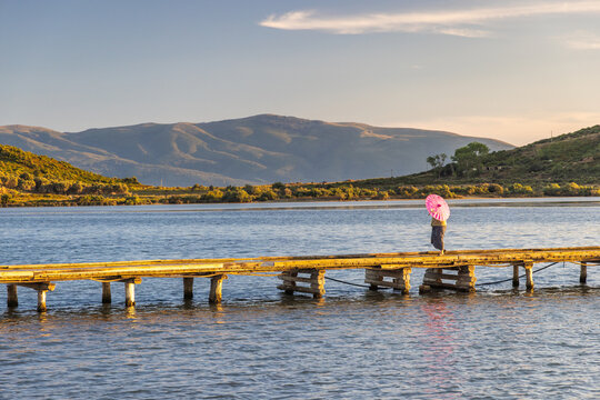 Seaside landscape near the city of Vlore in Albania, Europe. A person walks across a wooden bridge over calm water with a pink umbrella during a sunny day with mountain view