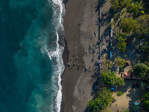 Aerial view of the Ngelarung Sea Offering Ceremony at Pura Segara Goa Lawah, with people in traditional attire on the black sand beach by the turquoise ocean in Dawan, Bali, Indonesia.