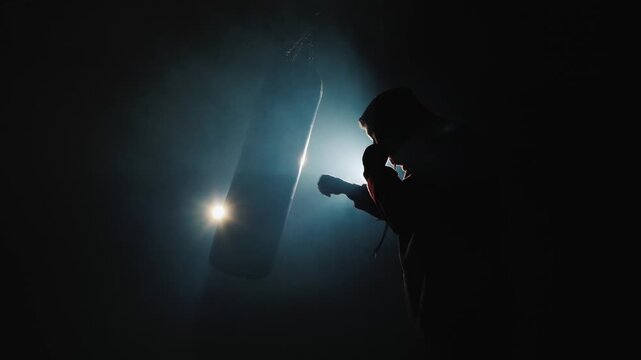 Silhouette of focused boxer training with heavy punching bag in dark smoky gym with cinematic lens flare.