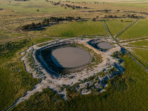 Aerial view of a large circular muddy water hole surrounded by green agricultural fields and sparse trees in the flat landscape of Hay, New South Wales, Australia.