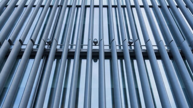 Vertical steel sunshade fins in front of a glass curtain wall with small facade spotlights on a mounting rail, low angle view in cool blue tones - solar shading and energy efficient building design