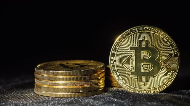 A stack of small shiny silver and gold coins next to a single large gold bitcoin as light slowly moves over them creating highlights on a black background.