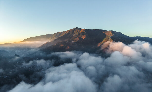 Aerial view of Dieng Plateau at sunrise, with volcanic mountains rising above a thick layer of clouds and soft golden light illuminating the peaks in Indonesia.