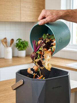 Person pouring organic food waste from a small green bin into a larger kitchen compost container.