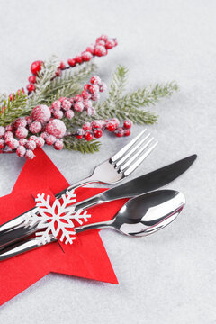 Holiday table setting with cutlery arranged in red star shaped cutlery holder decorated with white snowflake, showy Christmas tree branch on light gray textured background