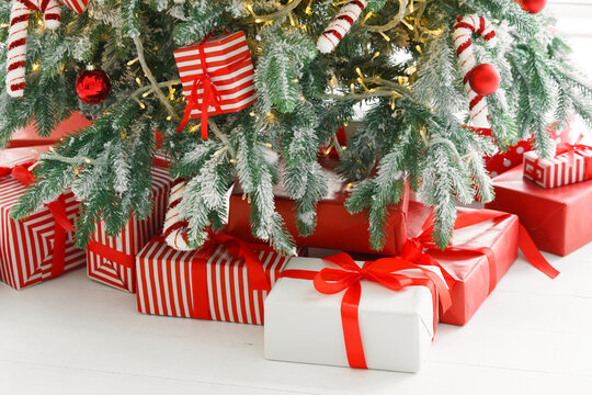 Beautiful presents, gift boxes wrapped in elegant red paper under green snowy festive Christmas tree with warm lights. Cozy winter holiday atmosphere perfect for seasonal celebrations and decor themes