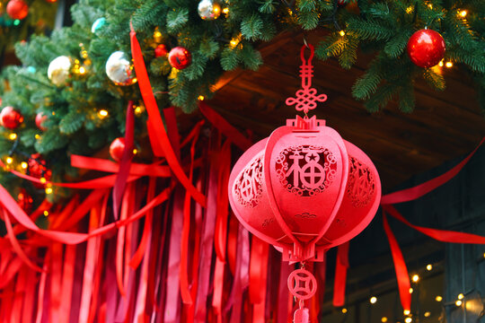 Festive Christmas decoration in Chinese inspired style with green evergreen garland, red ribbons, traditional lantern ornament and warm lights on a rustic wooden house. Holiday outside decor